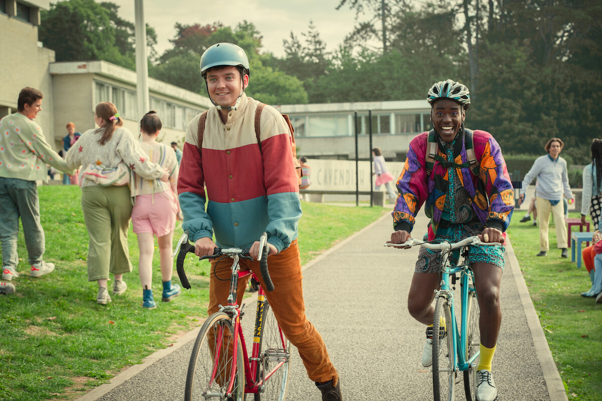 Asa Butterfield as Otis and Ncuti Gatwa as Eric ride their bikes outside of Cavendish College in Season 4 of ‘Sex Education.’