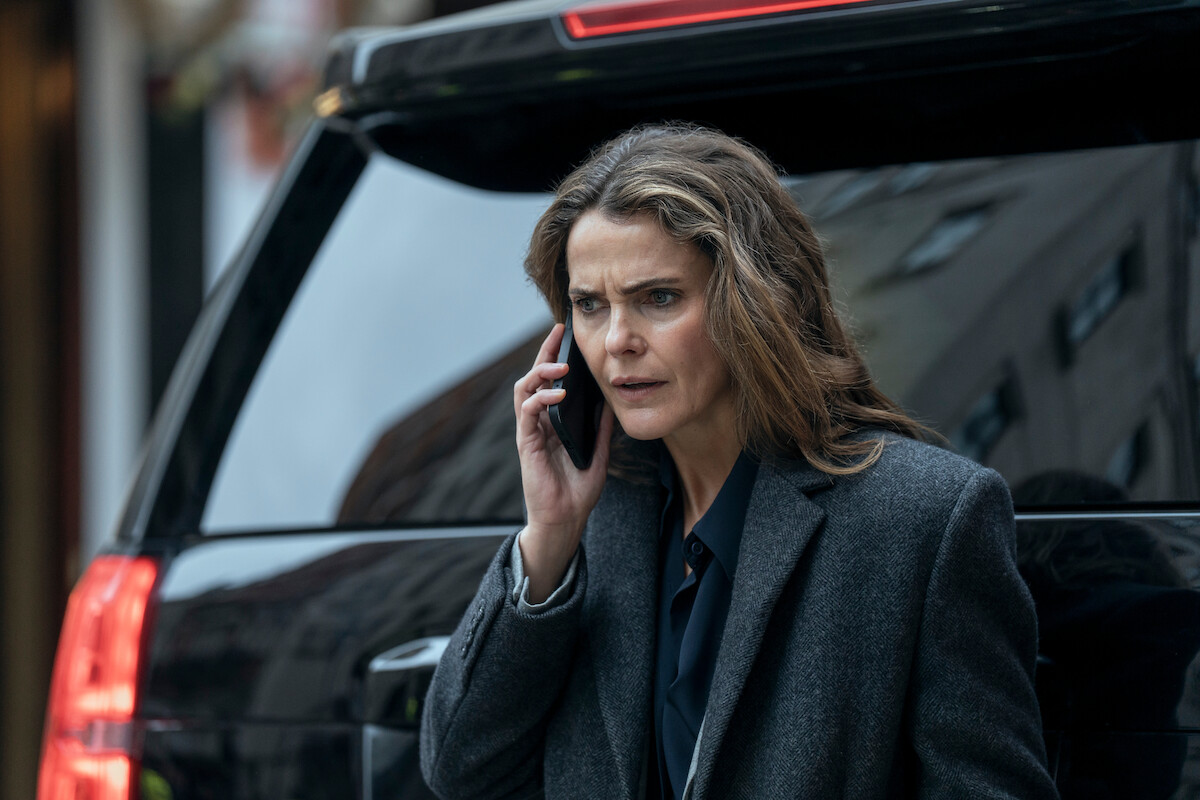 Woman in a gray coat standing beside a black SUV on a city street, holding a phone to her ear and looking concerned. Urban background with blurred buildings and natural daylight.