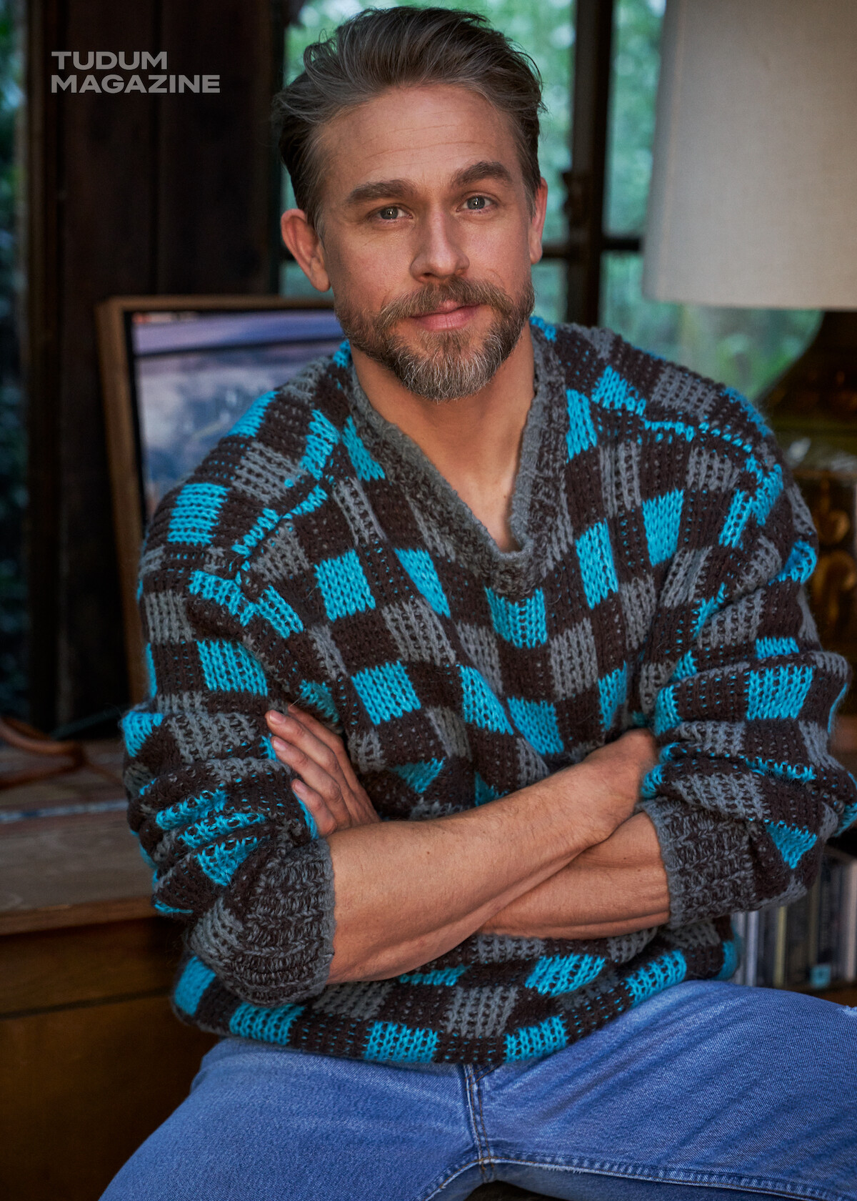 Charlie Hunnam sits on a stool in front of a credenza wearing a blue checked sweater and blue jeans.