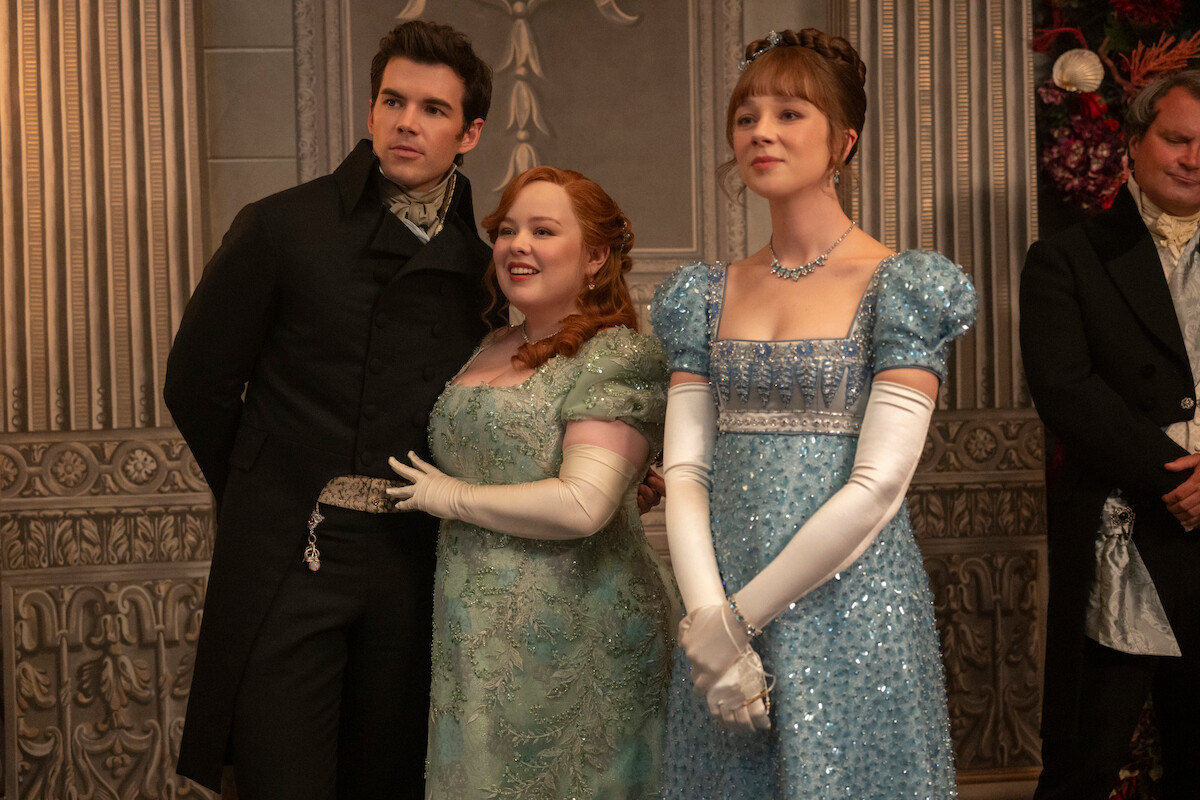Três pessoas trajando vestes elegantes da era regencial em um salão decorado com detalhes florais de pedra, sugerindo um baile ou encontro social formal.