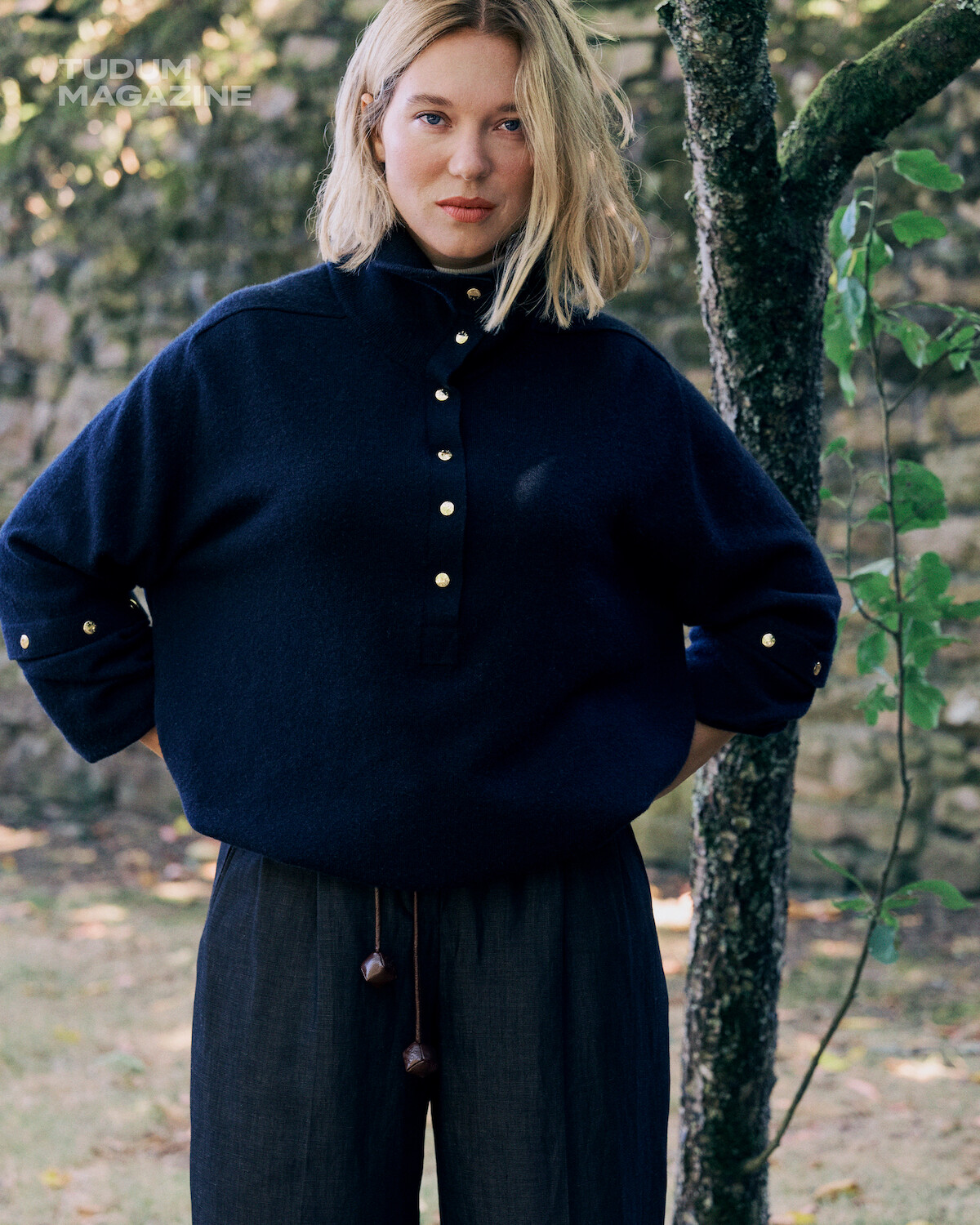 Woman with blonde hair in a navy sweater and dark pants stands outdoors in a grassy, shaded area with a stone wall and trees in the background, looking at the camera with a calm expression.