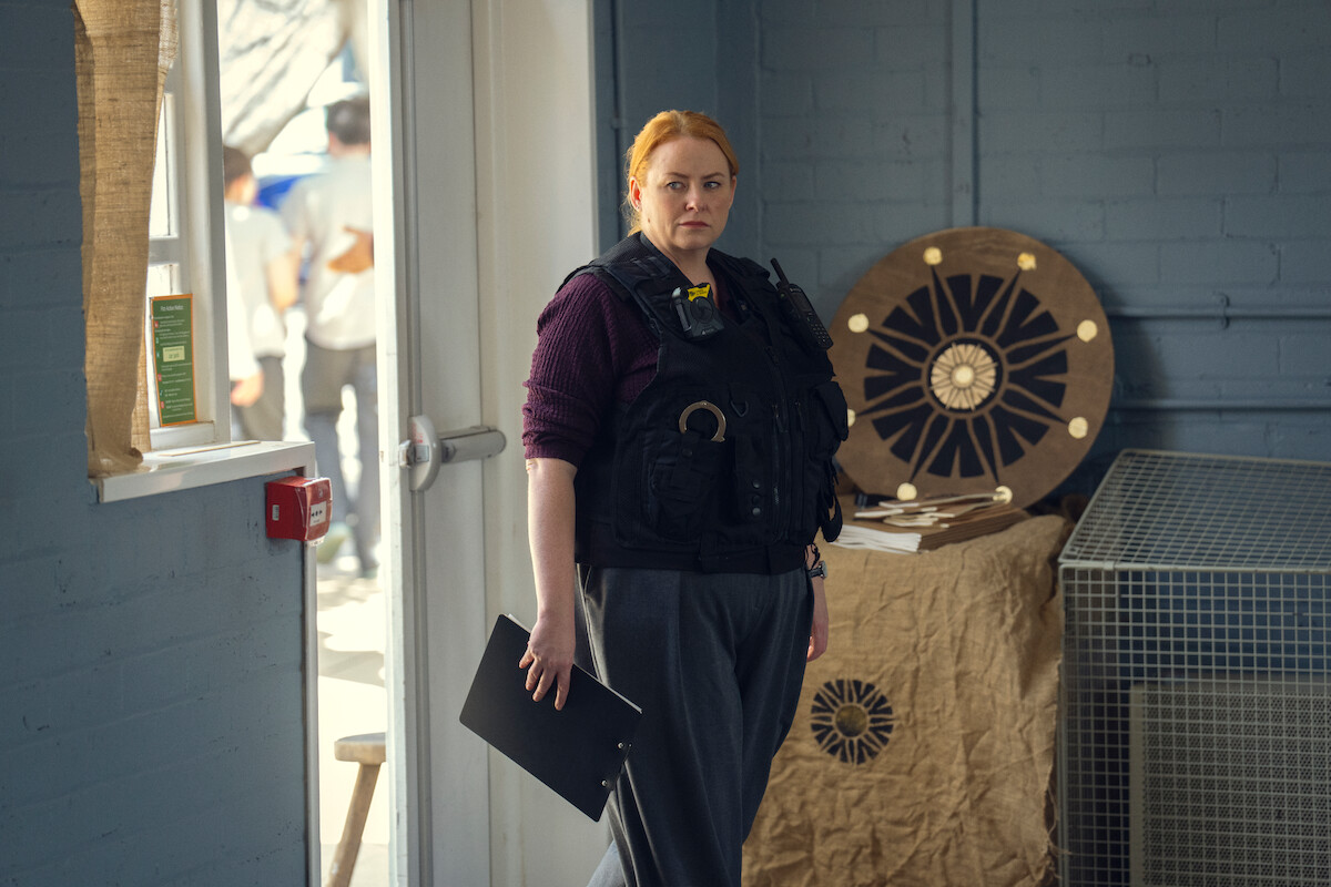 A woman in a tactical vest holds a clipboard, standing indoors by a door near a table with circular objects and a wire crate. The setting has blue brick walls and appears to be an office or institutional environment.