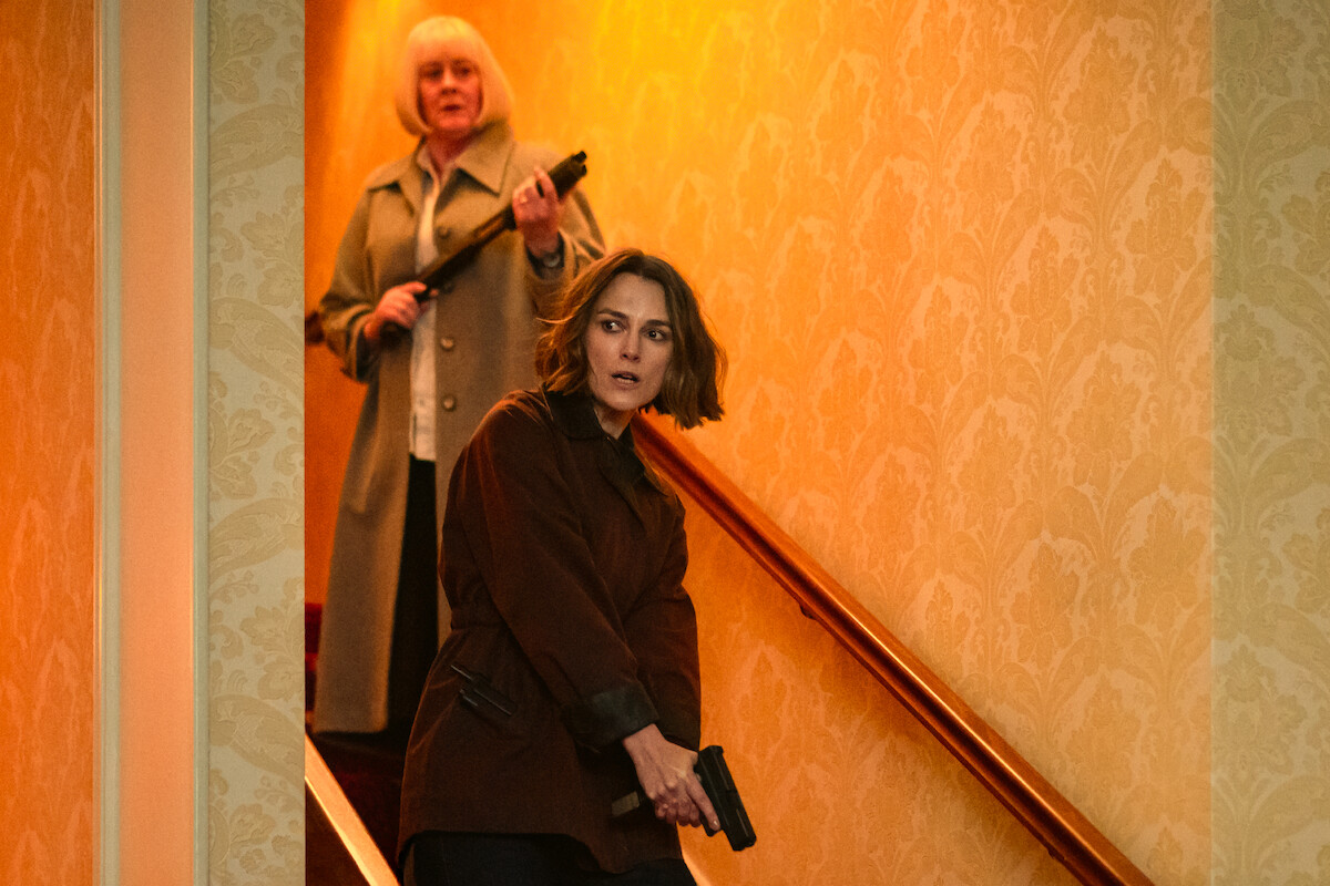 Two women cautiously descend a staircase in a warmly-lit hallway, one holding a handgun and the other a shotgun, both appearing alert and tense, wearing coats in an indoor residential setting.