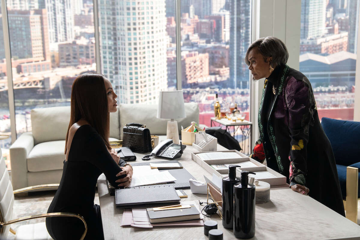 Two women in a modern high-rise office have a serious conversation across a desk, surrounded by paperwork and city views through large windows, creating a tense and professional mood.