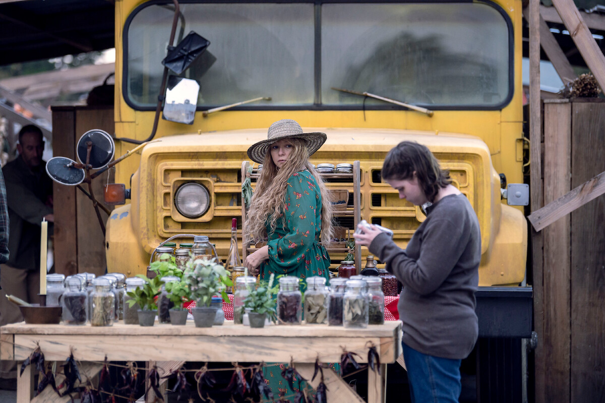 A group of settlers jar various plants and vegetables in front of a yellow school bus. 
