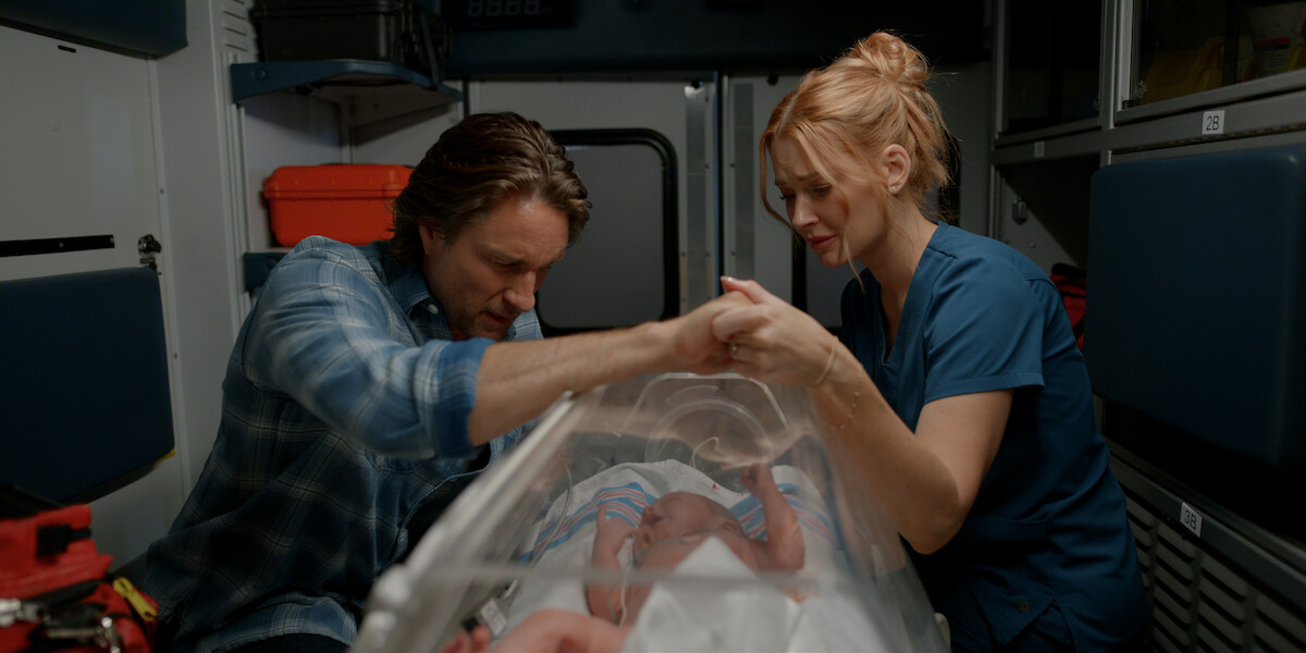 Two people care for a newborn baby inside an ambulance, focusing on the infant in an incubator, with medical equipment and supplies visible in the emergency vehicle setting.