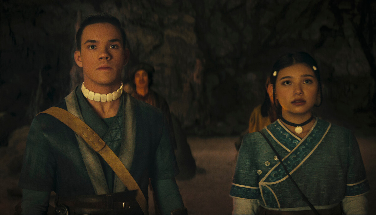 Two young people in blue traditional clothing stand in a dimly lit cave, looking ahead with serious expressions. Stone walls and another figure are visible in the background, suggesting a tense or significant moment.