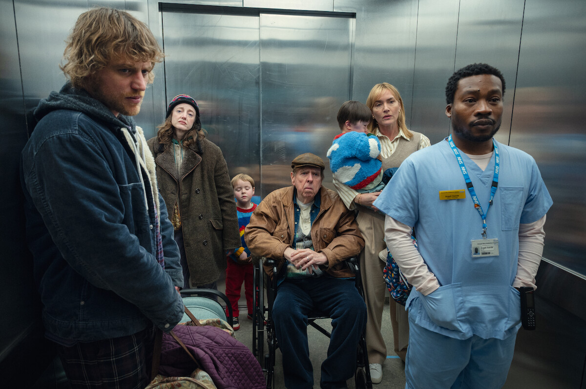 A group of people, including a nurse, a man in a wheelchair, children, and adults, stand together in an elevator with neutral or tense expressions.