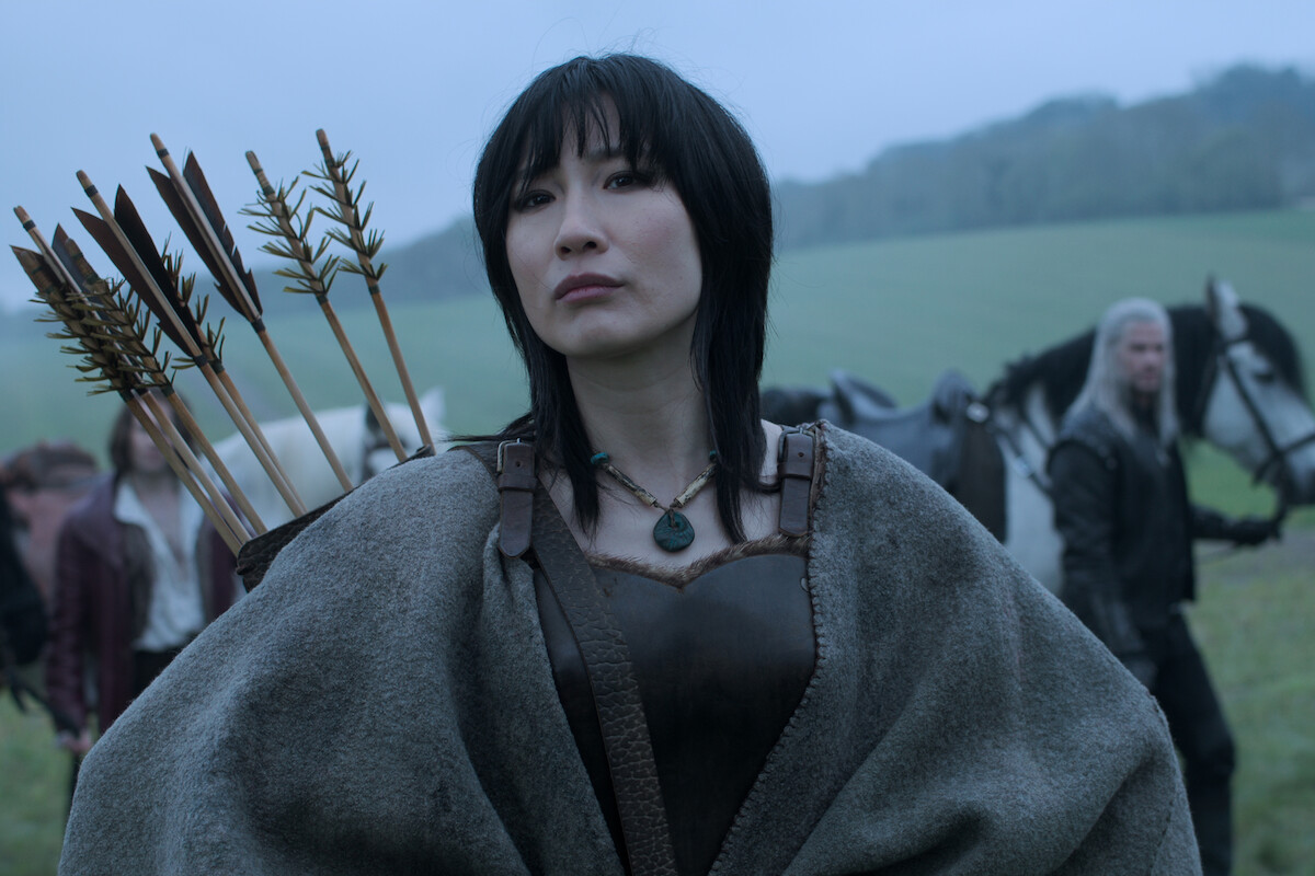 Confident woman archer with arrows stands outdoors in misty field, medieval fantasy setting, with other travelers and horses in the background, conveying a serious and determined mood.