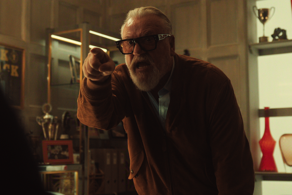 Older man with white hair and beard, wearing glasses and a brown jacket, angrily pointing in a modern office with trophies, shelving, and decorative items in the background.