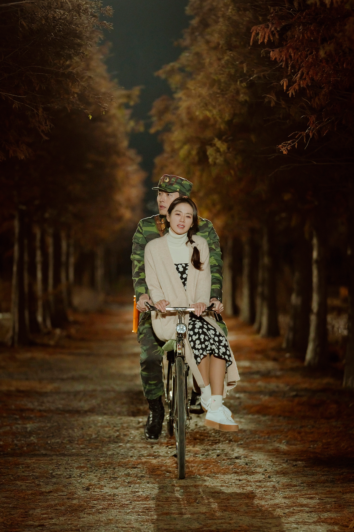 Um homem e uma mulher asiáticos dividem uma bicicleta em um caminho de floresta, com árvores dois dois lados da pista