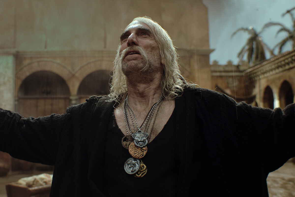 Older man with long hair and multiple medallions stands outdoors in a courtyard, arms raised, looking upwards as rain falls, evoking drama and intensity amidst stone arches and palm trees, with a somber, cinematic atmosphere.