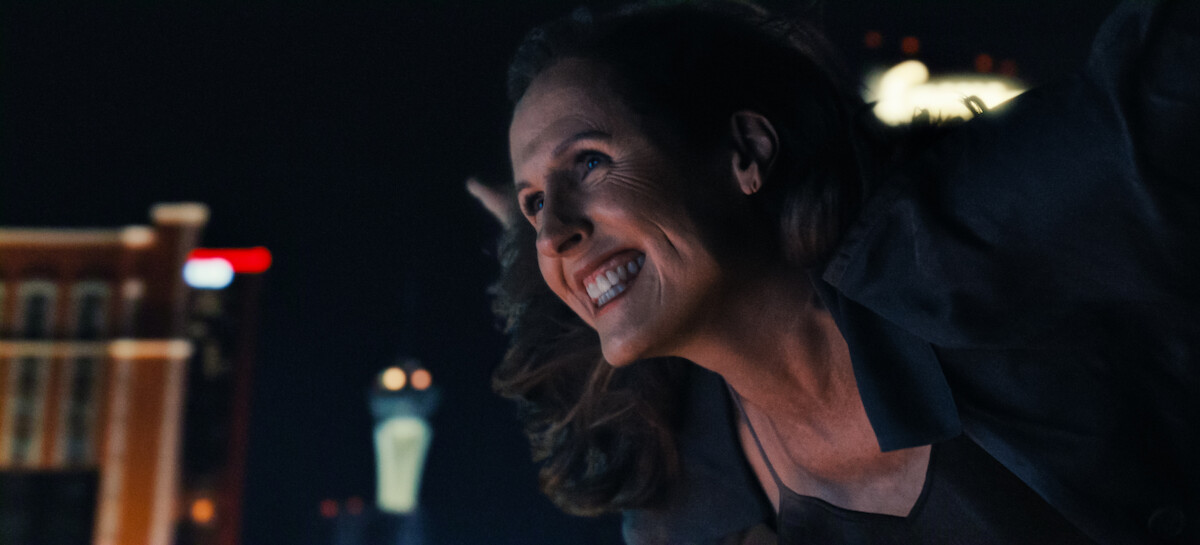 Smiling woman leaning forward at night, city lights and tall buildings in the background, urban environment with a joyful expression.