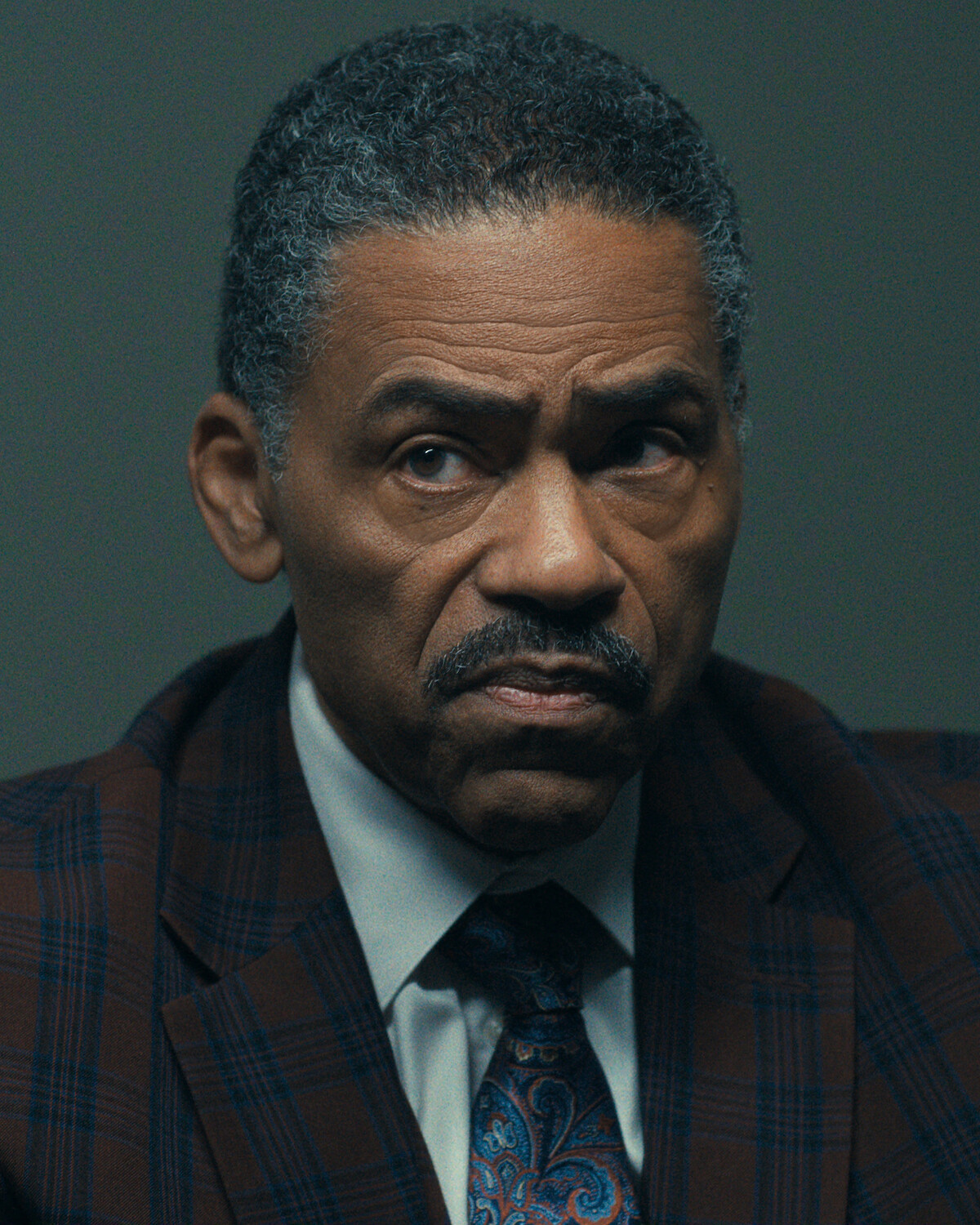 Middle-aged man in a plaid suit jacket, dress shirt, and paisley tie sits indoors under soft lighting, appearing serious and focused against a plain dark background.