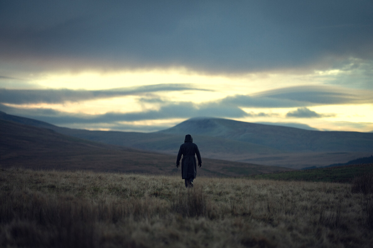 A solitary person in a dark coat walks across an open grassy field toward distant hills under a cloudy, moody sky at dusk. The landscape is vast and empty, evoking a sense of solitude and isolation.