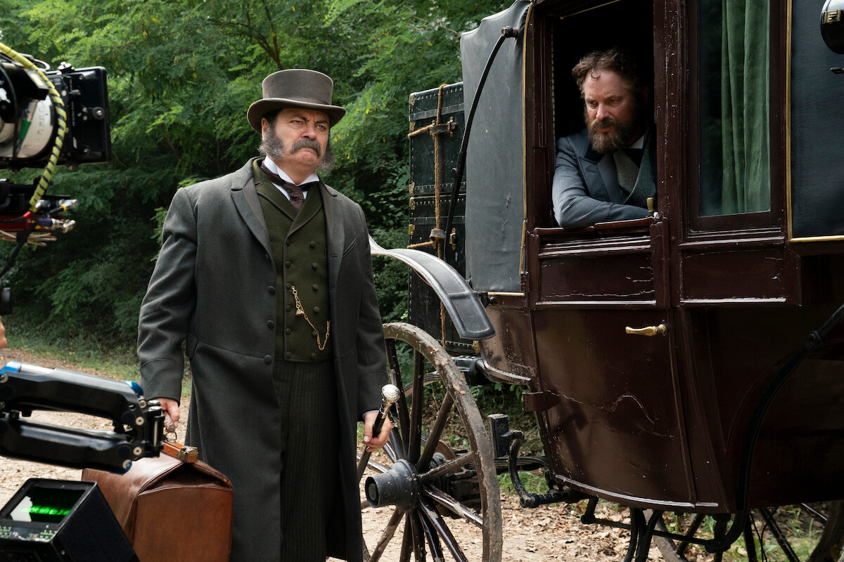 Two men in Victorian-era attire with a carriage; one stands holding a suitcase, and the other looks out from inside the carriage. Surrounded by greenery, the mood is tense and historical, with filming equipment visible.