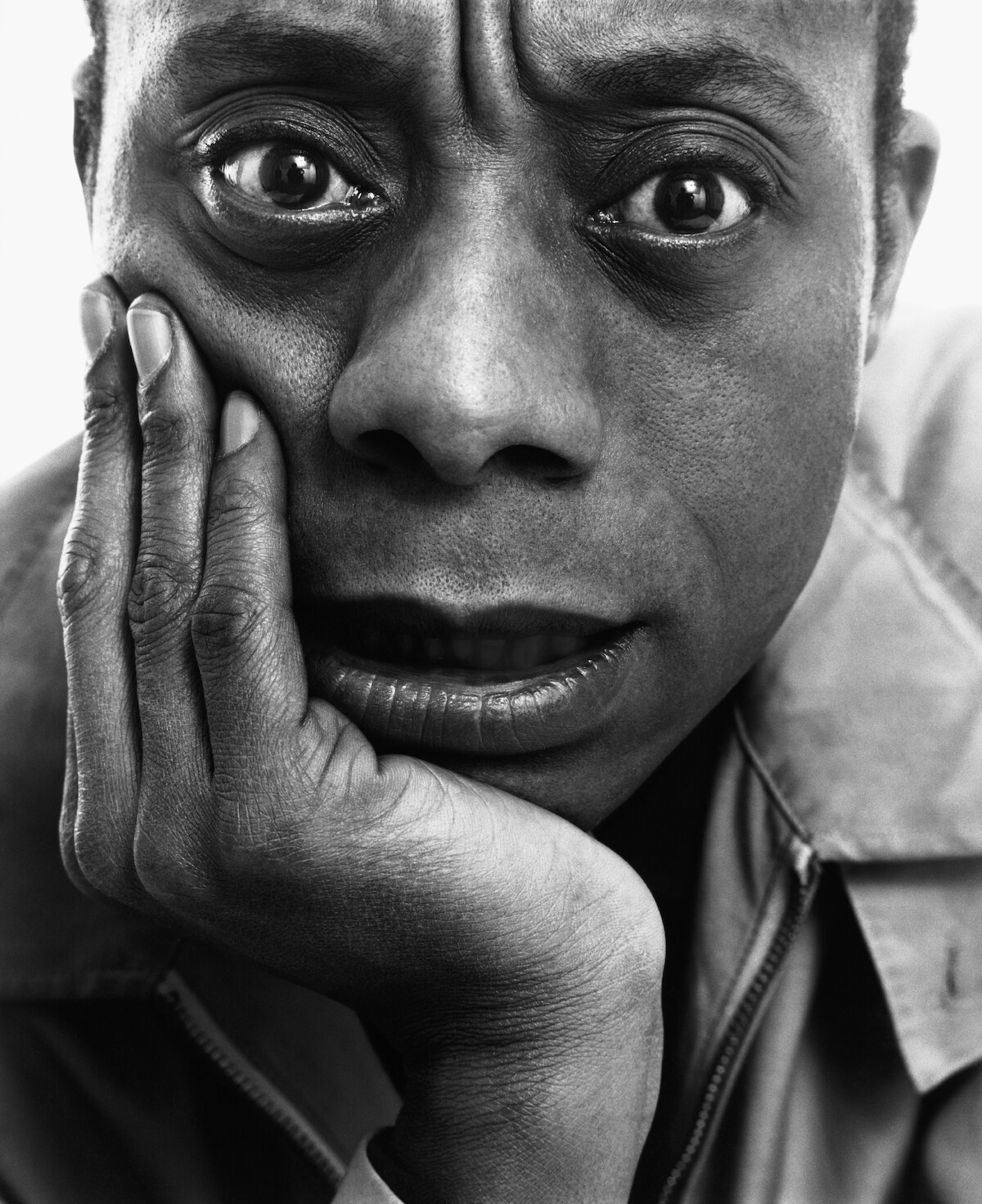 Black and white close-up portrait of a person resting their face on their hand, looking directly at the camera with an intense, contemplative expression, set against a plain background.