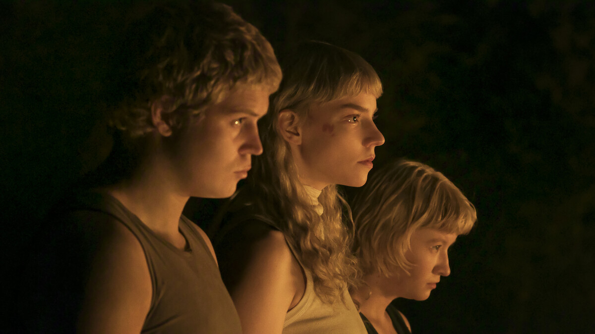 Three young people with blonde hair, lit by warm light, stand close together and gaze intently ahead in a dark, shadowy environment.
