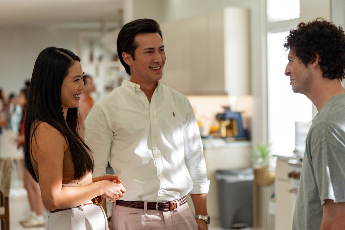 Three people standing and talking in a bright, modern kitchen during a social gathering, with other guests blurred in the background. The atmosphere appears relaxed and friendly.