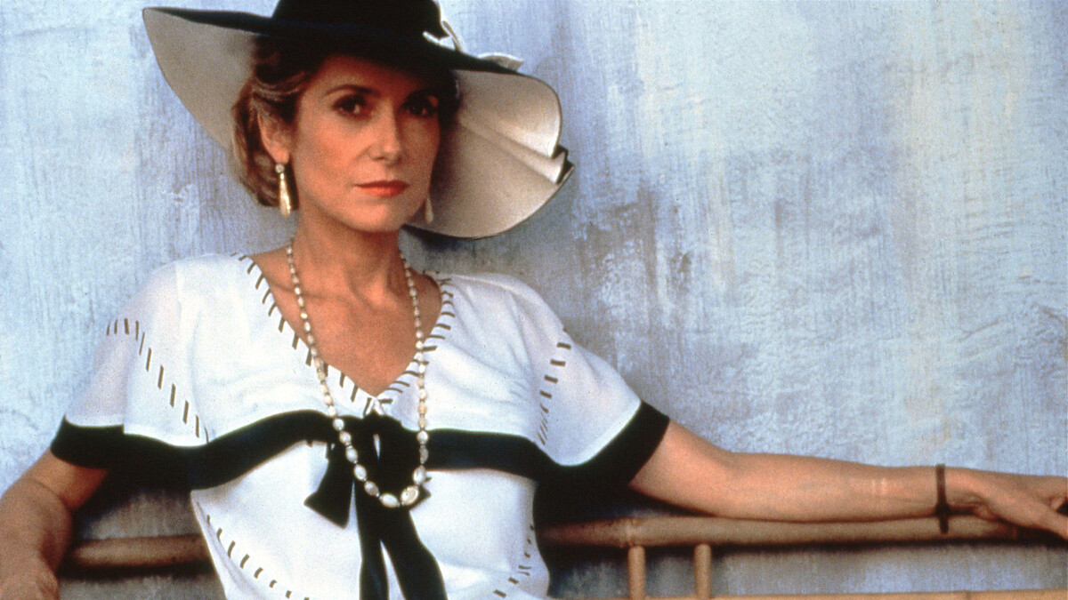 A woman in a white dress with black accents and a wide-brimmed hat sits against a textured, light-colored wall, wearing pearl necklaces and gold earrings, exuding a relaxed yet elegant demeanor.