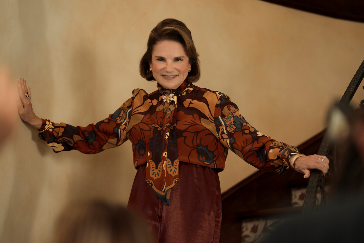 Older woman in a vintage floral blouse smiles, standing on a staircase indoors, hands on wall and railing, warmly engaging with people in a cozy home setting.