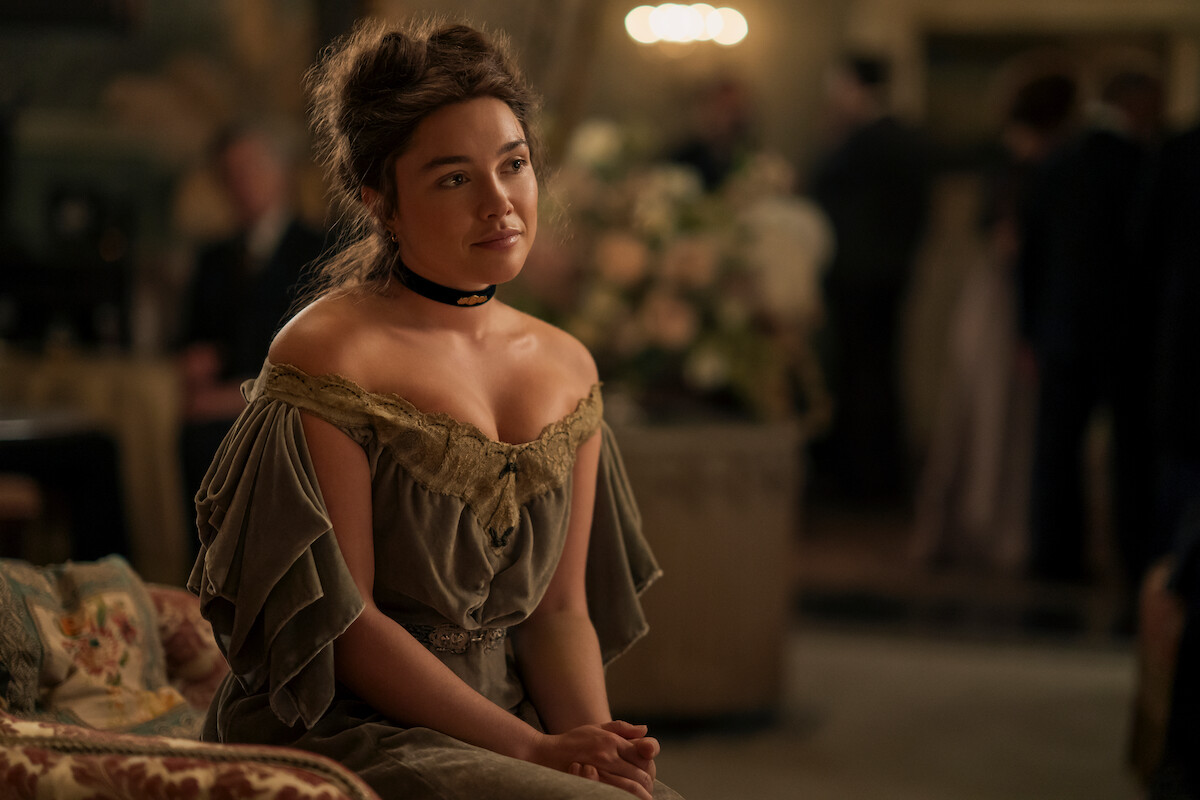 Mulher com um vestido vintage com os ombros à mostra sentada em um sofá estampado em uma sala elegante, com iluminação suave. Borrado ao fundo, há pessoas e arranjos de flores, sugerindo uma reunião formal ou em ambiente histórico.