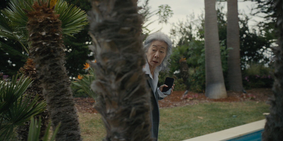 Older woman holding a phone stands in a garden near a pool, partially hidden behind palm trees, looking surprised or cautious. Lush greenery and tall trees in the background.