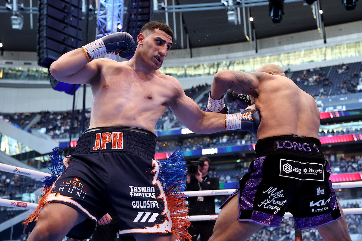 Two boxers in a boxing ring during a match, one delivering a punch to the other. They are surrounded by a large indoor stadium filled with spectators. Bright lights and digital screens are visible in the background.