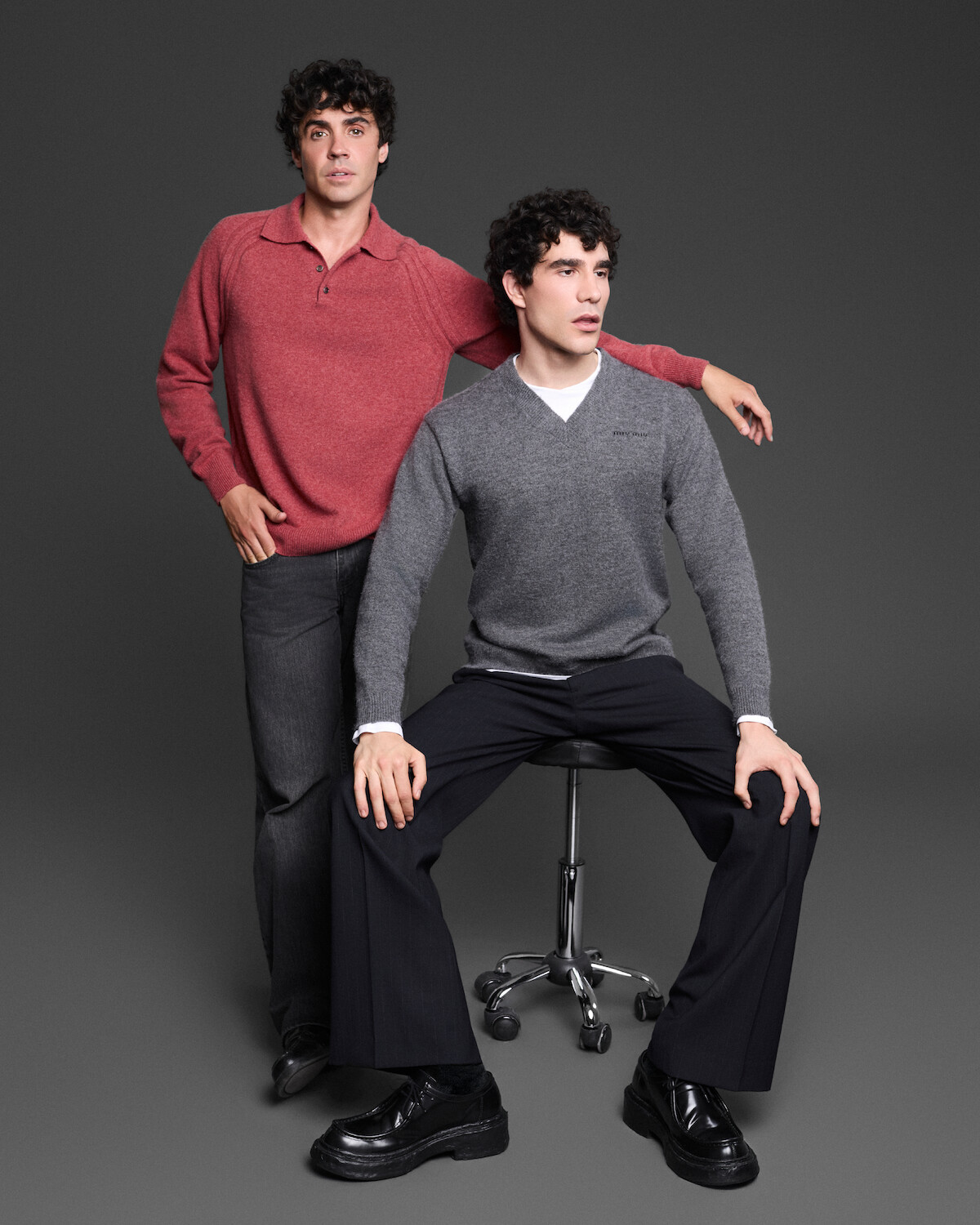 Two men pose in a studio with a dark background; one stands wearing a red sweater and jeans, while the other, dressed in a gray sweater and black pants, sits on a stool.