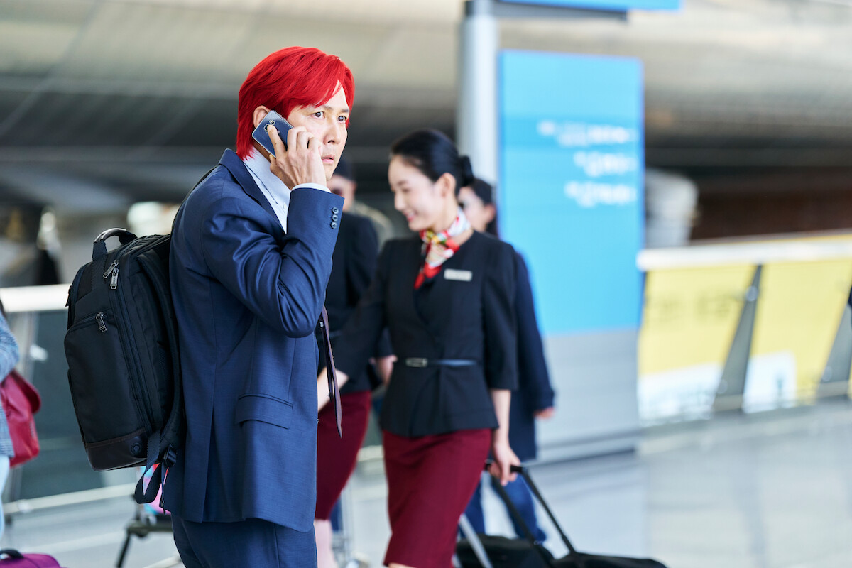 Lee Jung-jae as Seong Gi-hun with bright red hair in a blue suit talking on the phone at the airport, with airline staff in the background in ‘Squid Game’ Season 2.