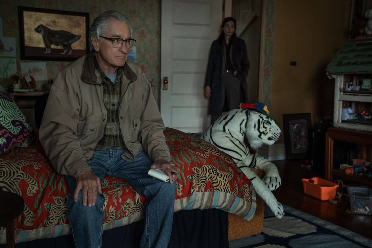 Older man sits on patterned bed holding remote in cozy, cluttered room with wallpaper, stuffed white tiger toy, and woman standing by the door in background. Warm lighting and eclectic decor give a homey feel.