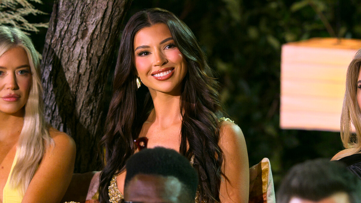 Woman with long dark hair smiling, seated outdoors at a nighttime event under warm lighting, surrounded by other people and greenery, creating a lively and elegant atmosphere.
