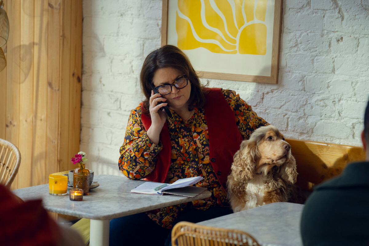 Woman in floral shirt, glasses, and red vest sits in cozy cafe with notebook, talking on phone, accompanied by a dog on the bench. Warm, relaxed atmosphere with candles and modern art on the wall.
