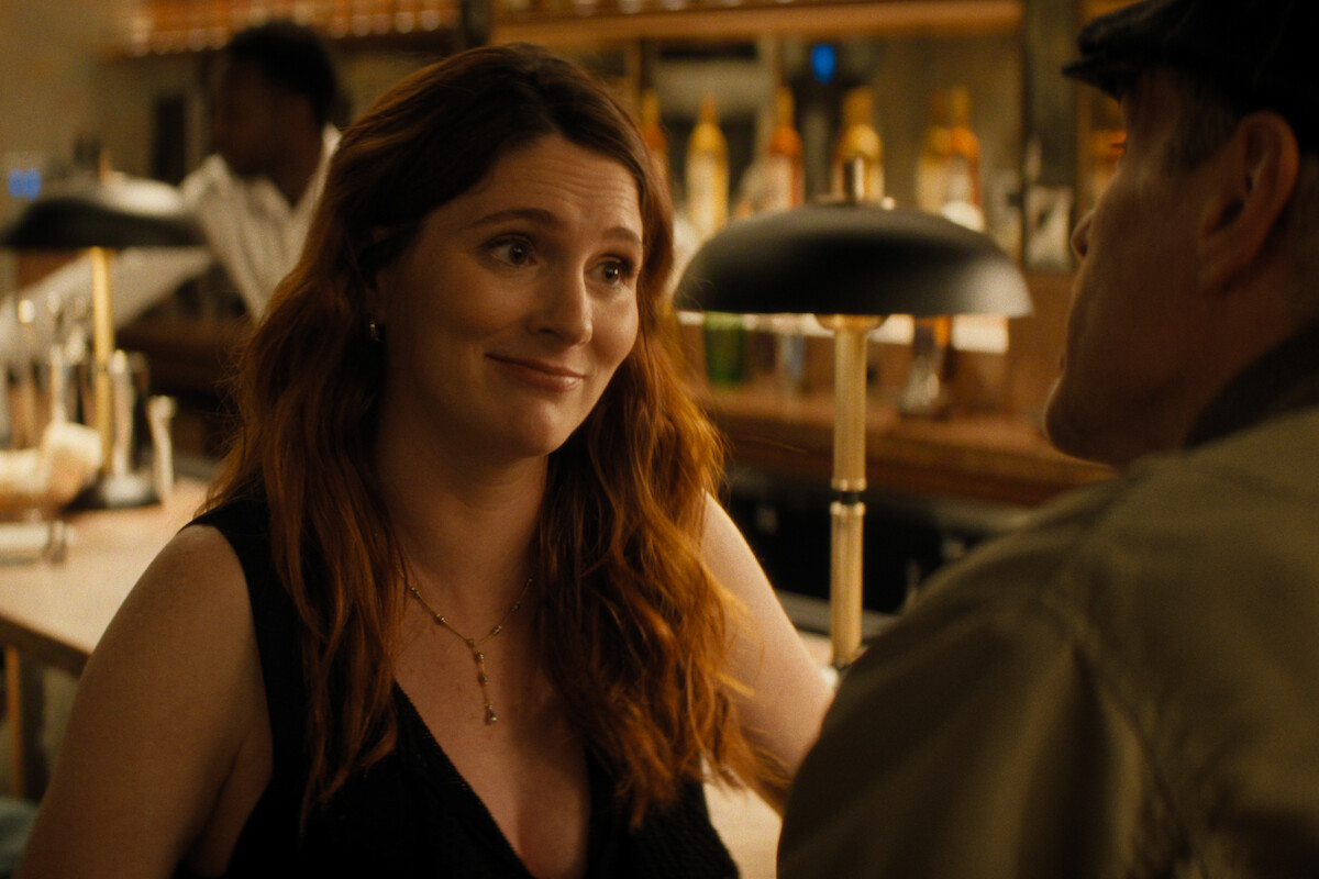 A woman with long brown hair sits at a bar, smiling and talking to a man in a hat. The background shows bottles, a bartender, and warm lighting, creating a cozy atmosphere.