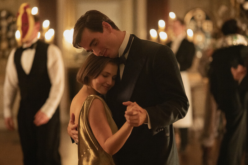 A couple in elegant attire dance closely at a formal, candlelit party, surrounded by other guests in masks and tuxedos, creating an intimate and romantic atmosphere.
