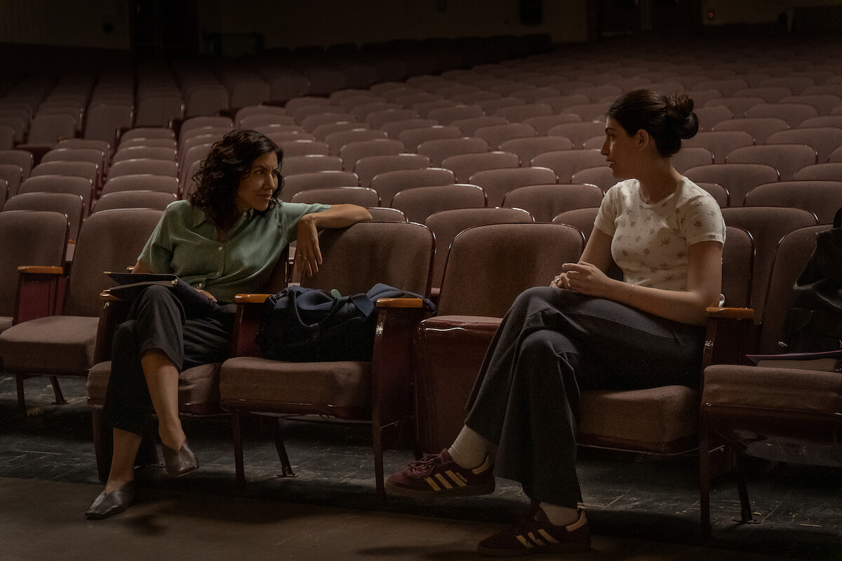Two women sitting and talking in an empty theater auditorium, surrounded by rows of vacant seats, with bags on the chairs, suggesting a quiet discussion or meeting in a dimly lit, spacious environment.