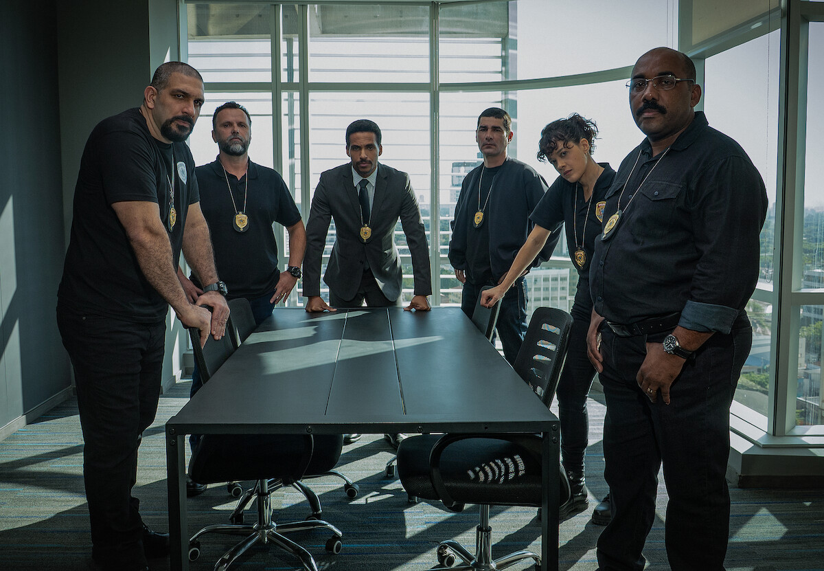 Grupo de 5 homens e 1 mulher, com distintivos no pescoço, se reúne em volta de uma mesa de delegacia policial
