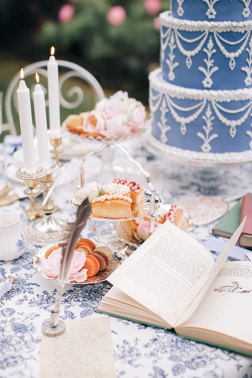 Elegant table set for tea outdoors with pastries, candles, open book, feather pen, and a large blue tiered cake, all creating a romantic, vintage, and whimsical atmosphere.
