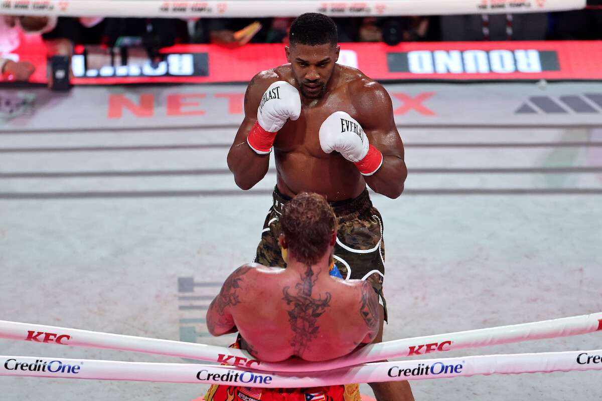 Jake Paul on the ropes as Anthony Joshua stands in front of him.