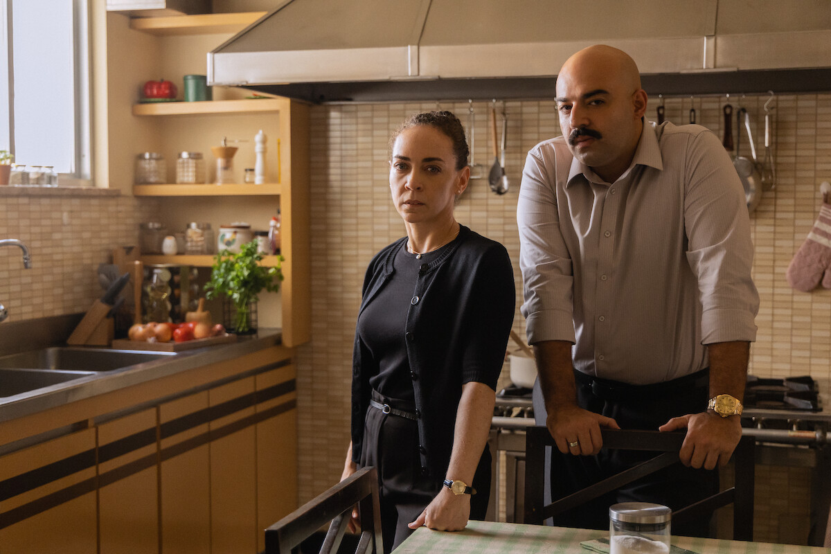 Um homem careca e de bigode usa roupa social em uma cozinha