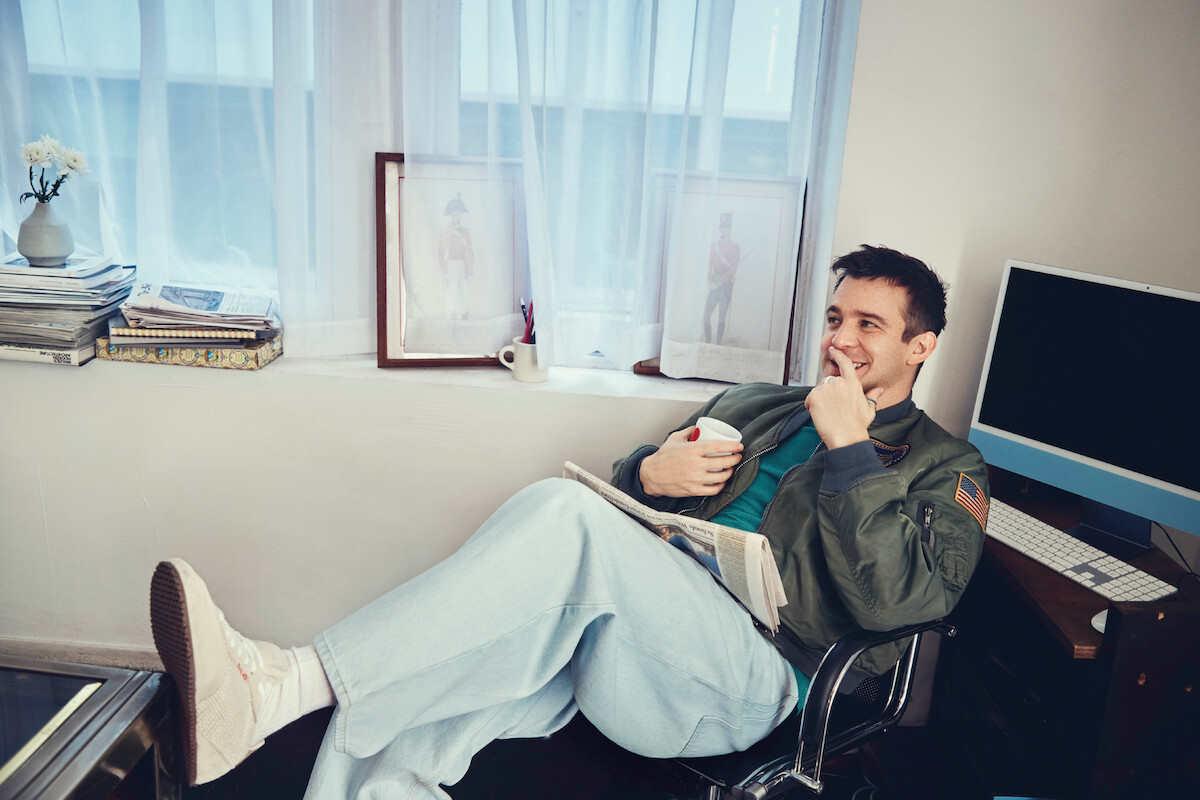 Um homem relaxa sentado em uma cadeira de escritório com os pés apoiados na mesa, segurando uma xícara de café e sorrindo. Ao redor, há uma mesa com computador, livros, quadros emoldurados e flores ao lado de uma janela com cortinas leves, compondo um espaço de trabalho casual e acolhedor.