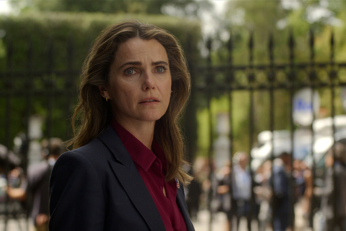 Woman with long brown hair in a dark blazer and burgundy shirt stands outdoors in front of an iron fence, with a crowd and vehicles blurred in the background, appearing concerned or thoughtful.
