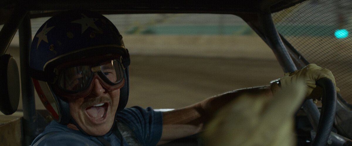 Man wearing a helmet and goggles smiles excitedly while driving a race car on a dirt track, visible through the vehicle’s interior, suggesting a lively racing atmosphere.
