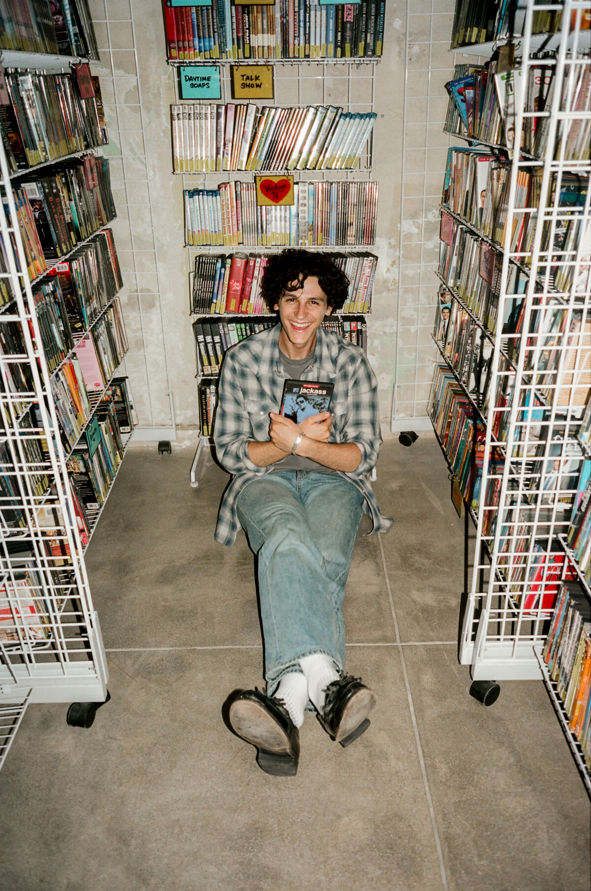 Owen Painter sitting on the floor in a narrow video store aisle holding DVDs and smiling.