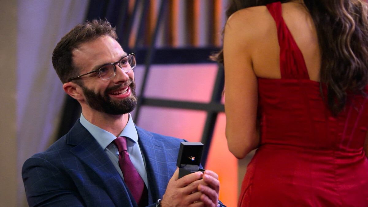 Man in a suit kneeling and proposing with a ring to a woman in a red dress indoors, both smiling in a warmly lit room.