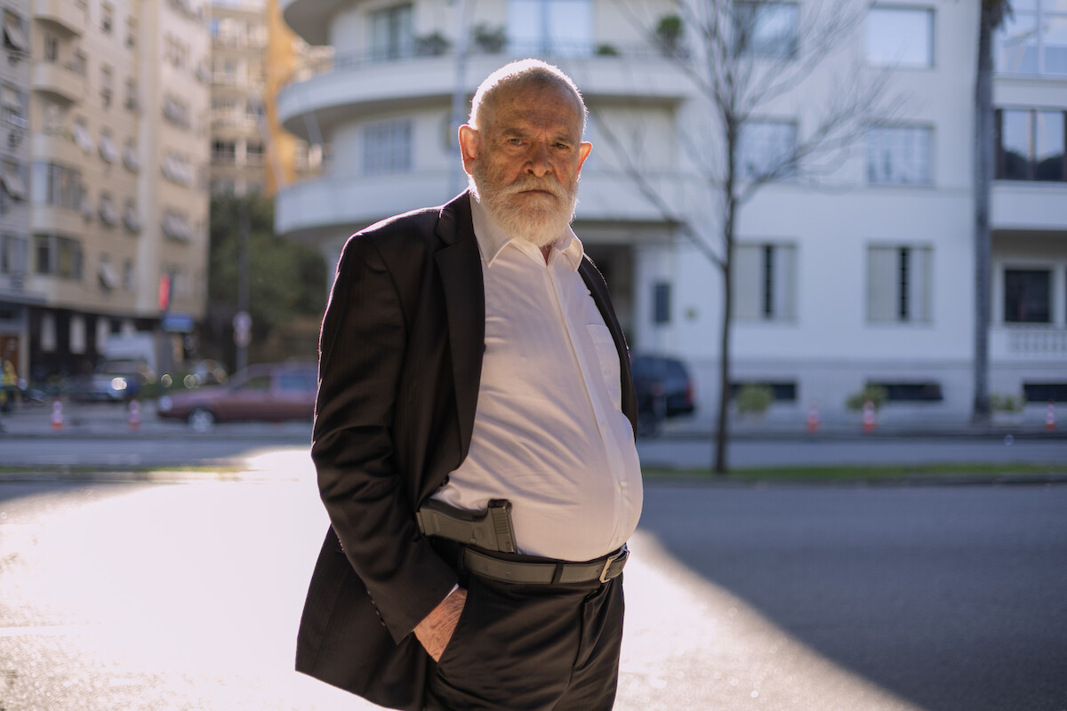 Um homem de cabelos e barba brancos veste camisa branca e terno preto. Ele tem uma arma presa junto ao cinto