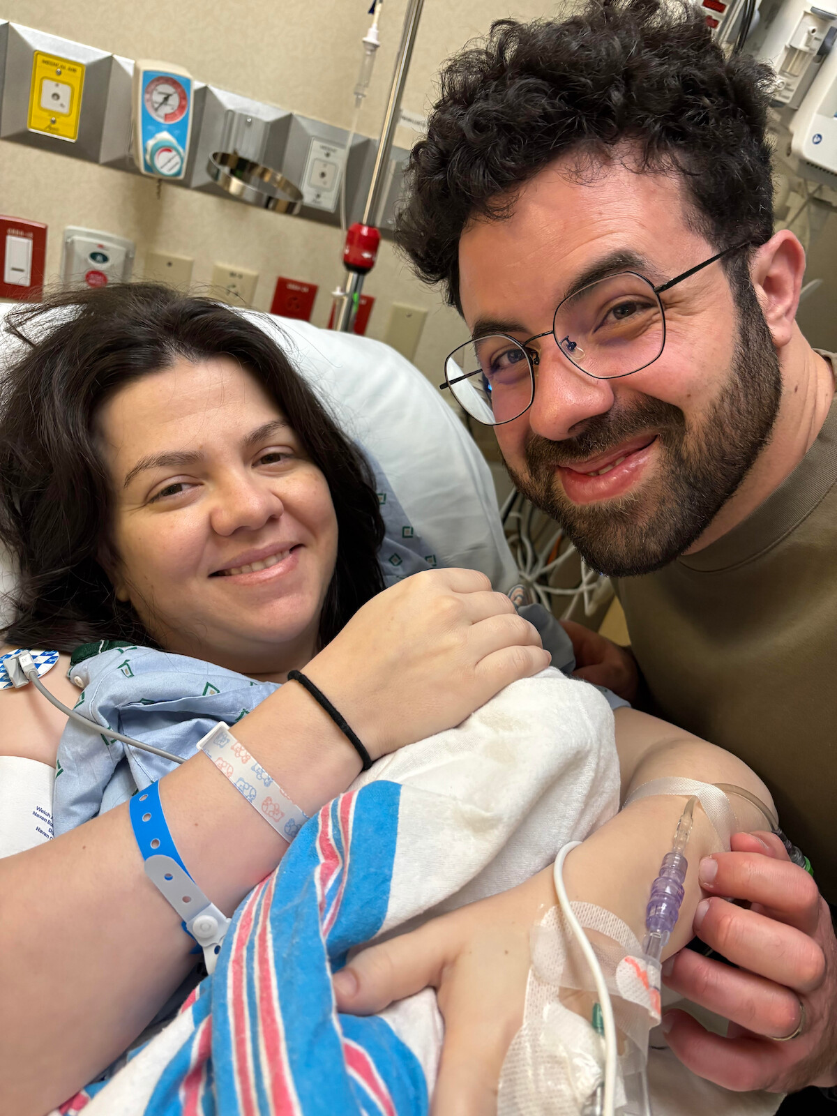Smiling woman in a hospital bed holding a newborn baby with medical wristbands, next to a man who is smiling. Hospital equipment and beige walls visible in the background.