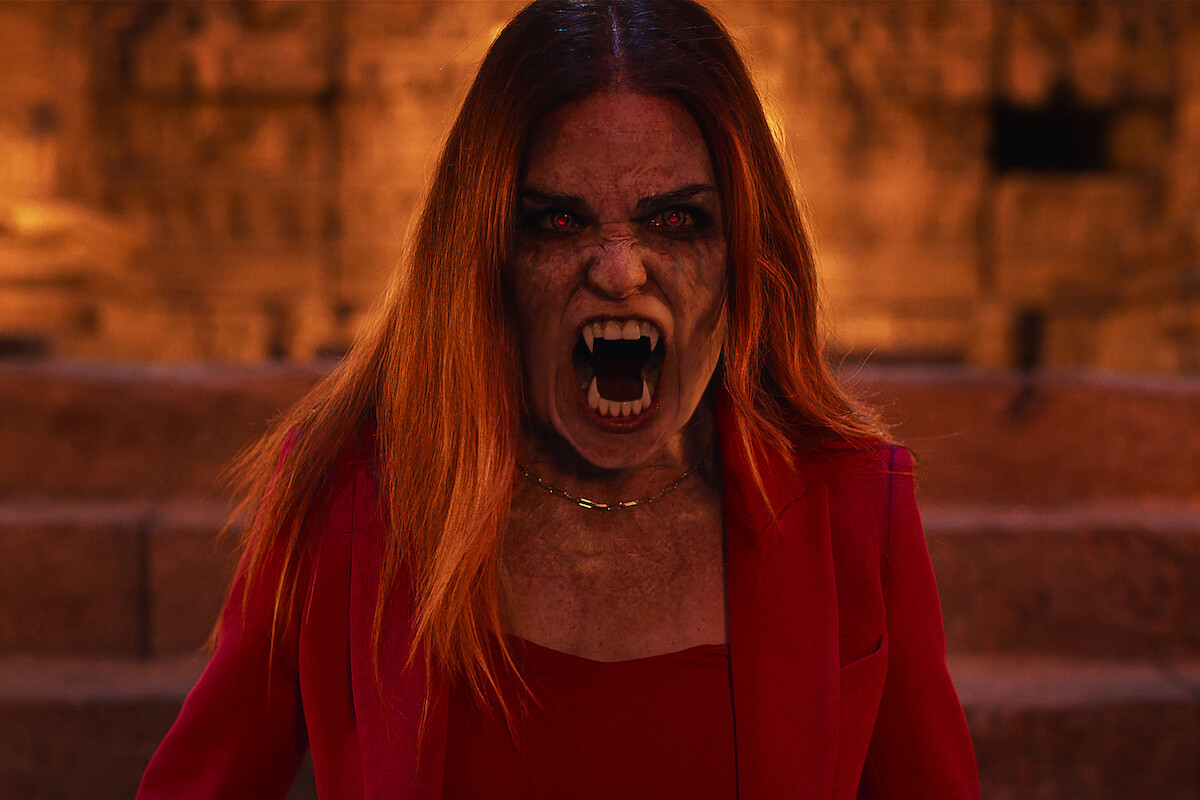 Woman with vampire fangs and glowing eyes in a red outfit snarling fiercely, standing in front of stone steps and a warm, dimly lit background.