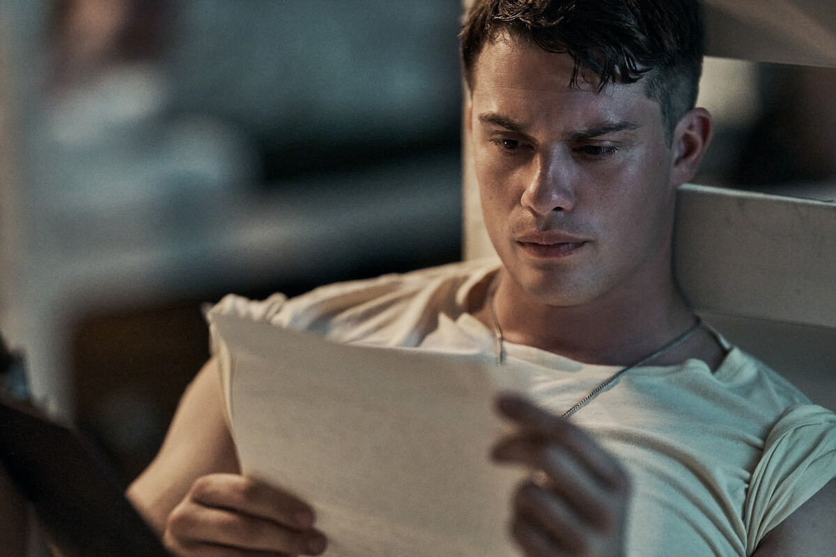 Nicholas Galitzine as John McLaughry in a casual t-shirt sitting indoors, leaning against a wall or bed frame, reading a letter with a serious expression, background softly blurred, conveying a contemplative or emotional moment.