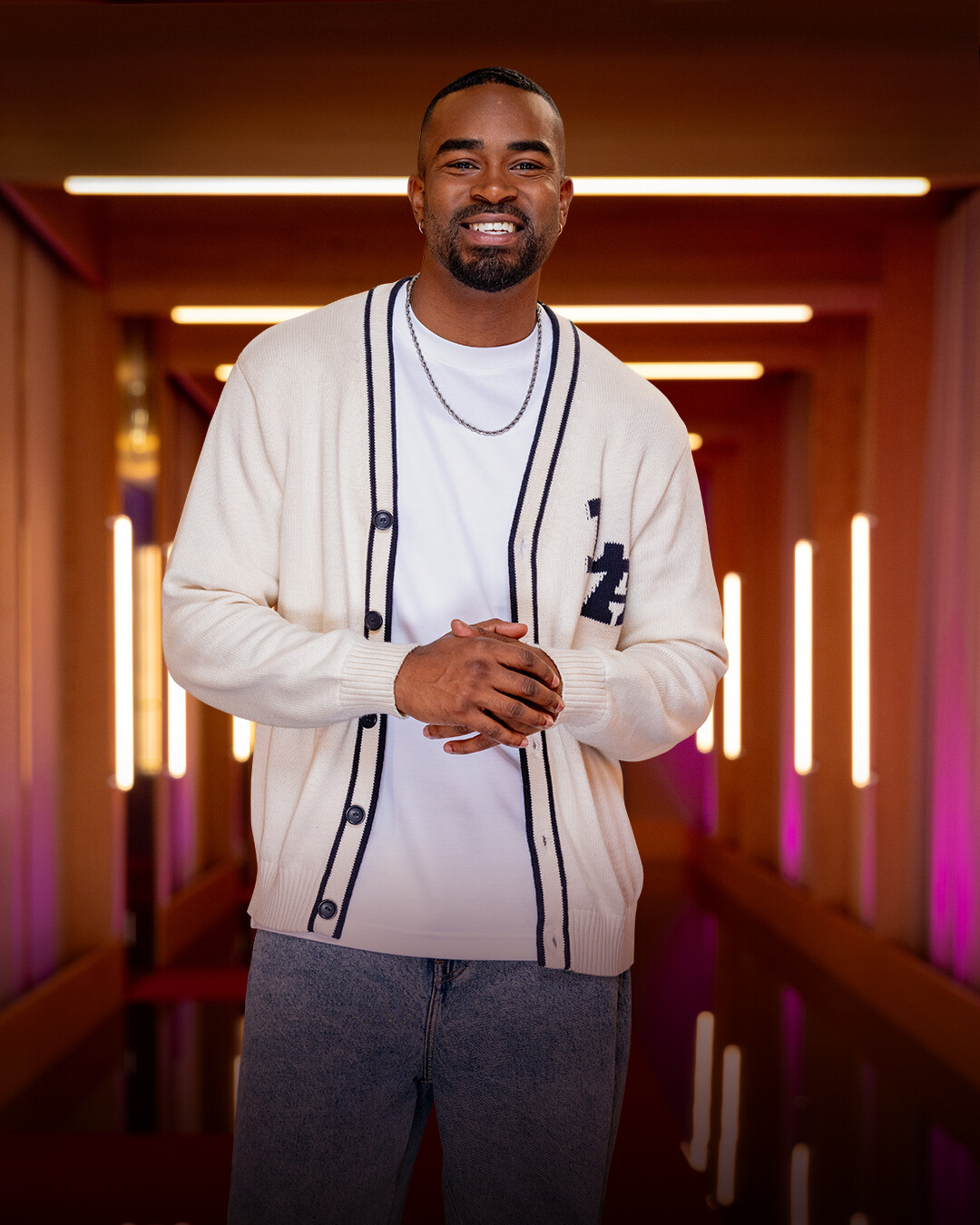 Smiling man wearing a white cardigan and casual jeans stands in a modern, warmly lit hallway with vertical lights and soft ambient colors in the background.